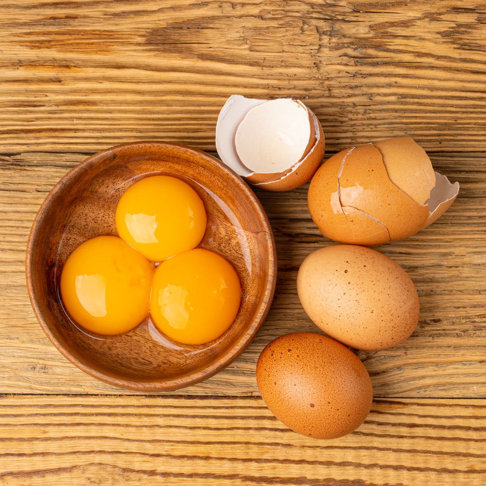 A wooden bowl with three eggs in it with their shells next to the bowl
